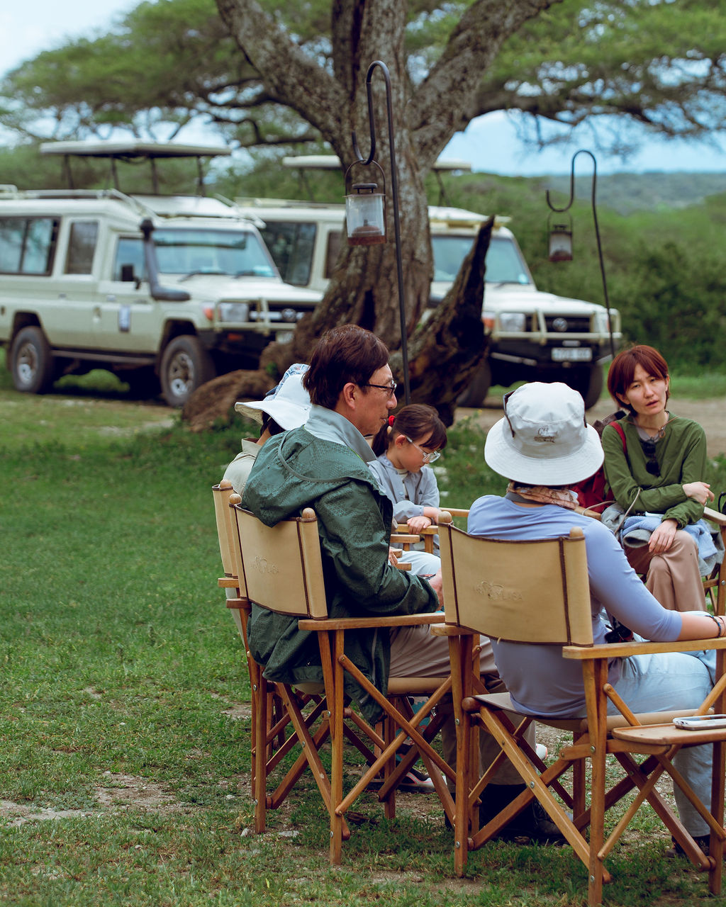 Guests relaxing in camp chairs with the safari vehicles behind